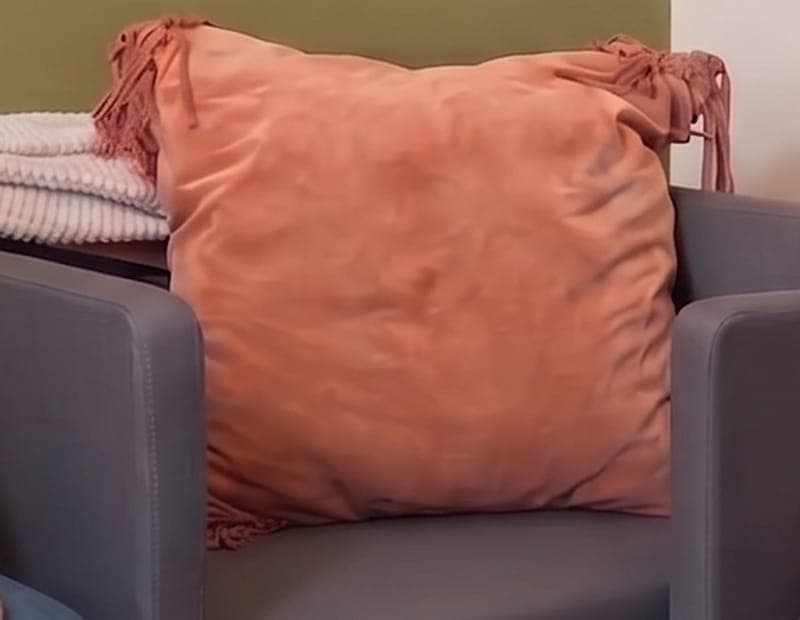 A large terracotta cushion with fringed corners on a gray armchair, a white ribbed throw folded beside it.