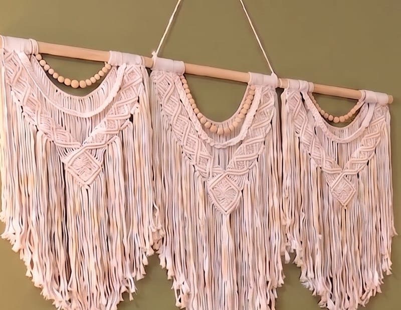 A three-panel cream macramé hanging on a wooden dowel against an olive green wall.
